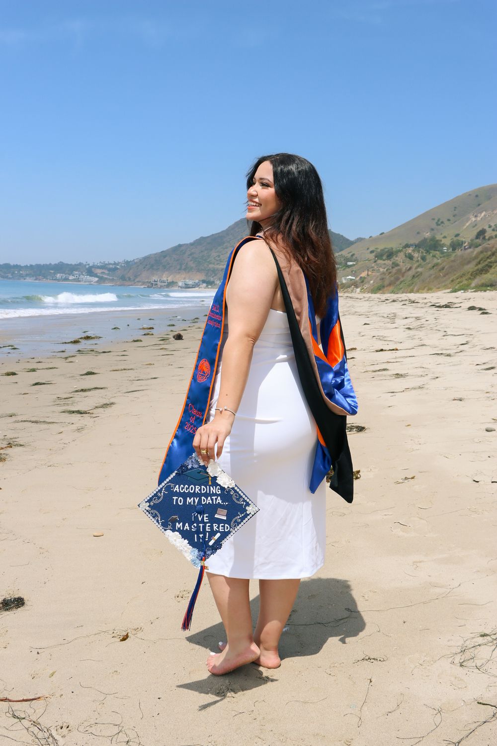 Graduate poses with diploma on a scenic beach with crashing waves in the background.