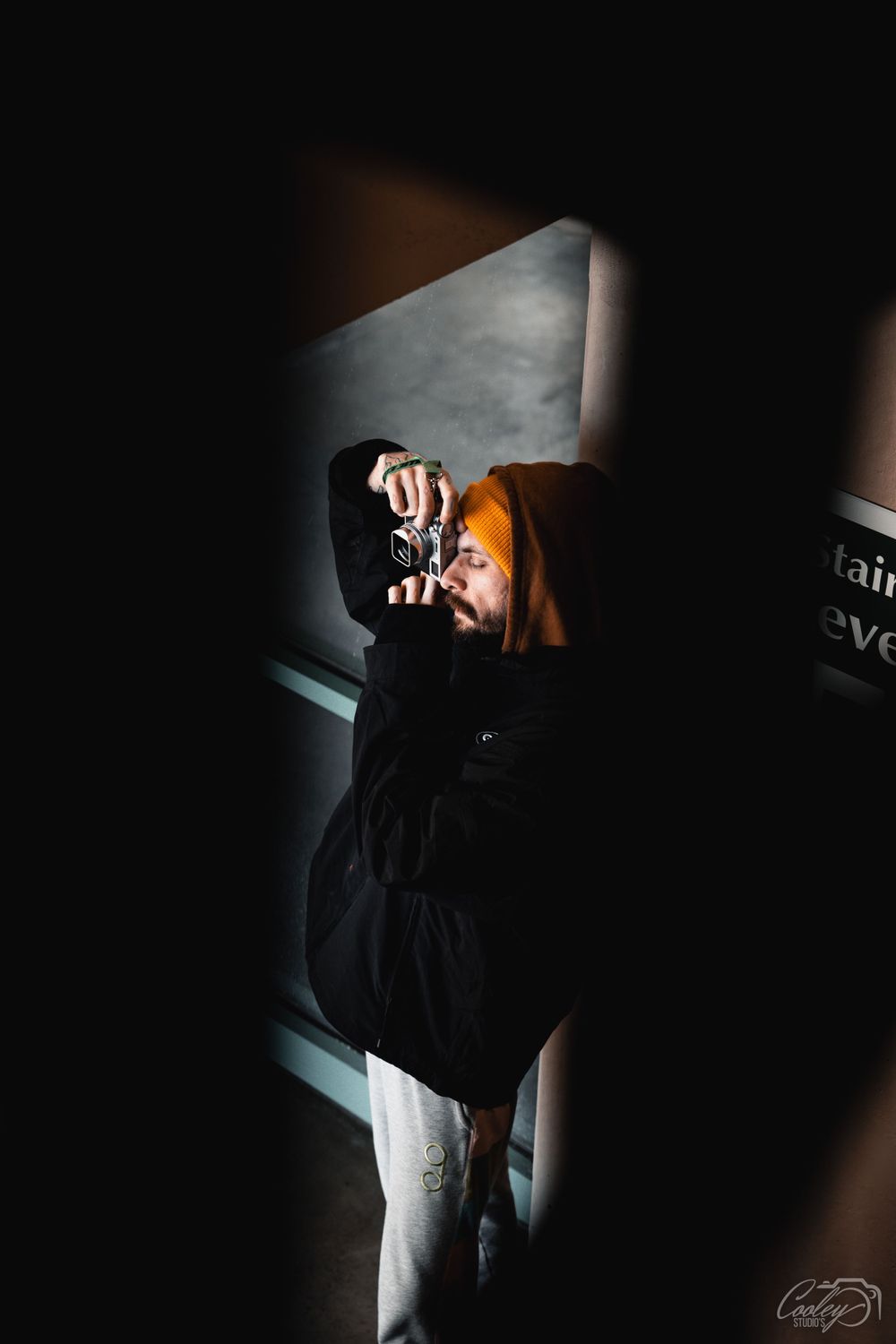 Photographer in orange beanie and black jacket shooting with vintage camera in dramatic light.