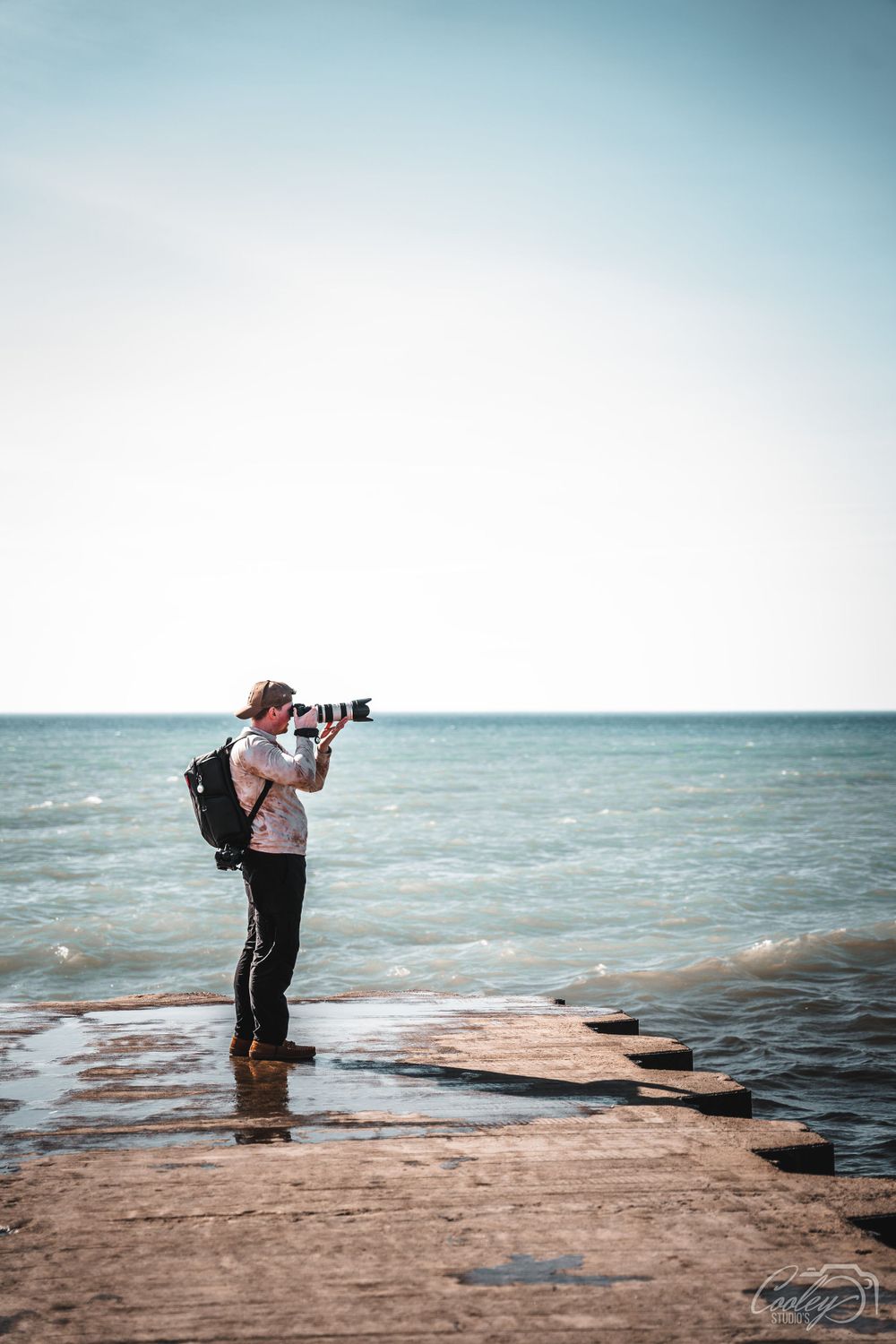 Photographer with backpack shooting ocean horizon while standing on stone pier at beach.