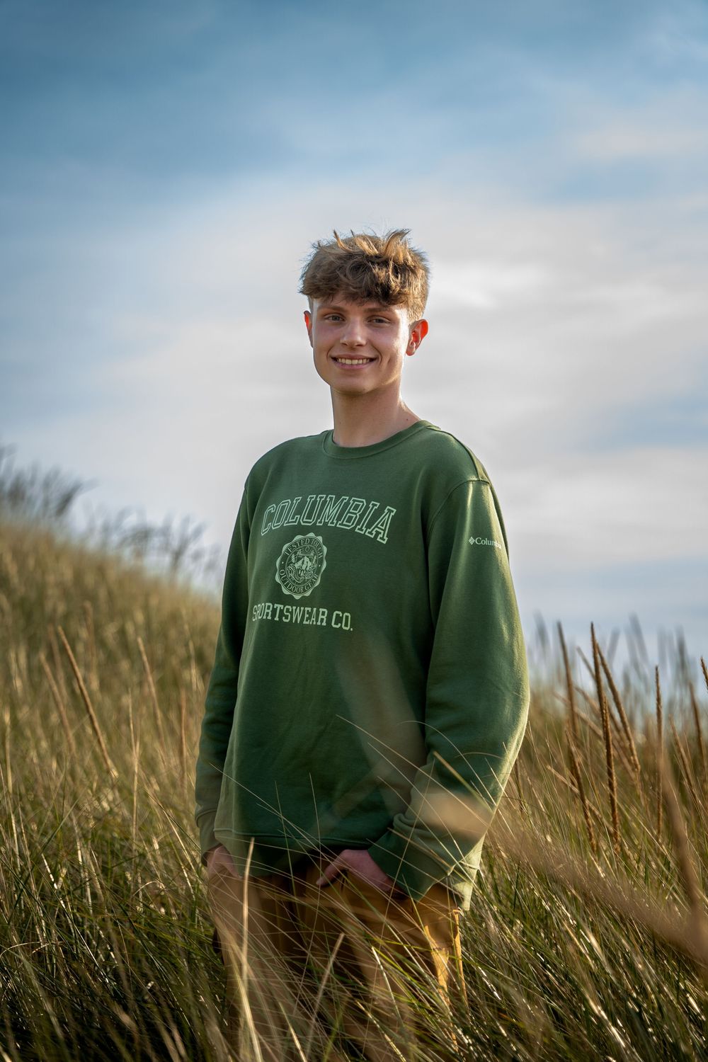 Teenage boy smiling in green Columbia Sportswear sweatshirt standing among tall golden grass outdoors.