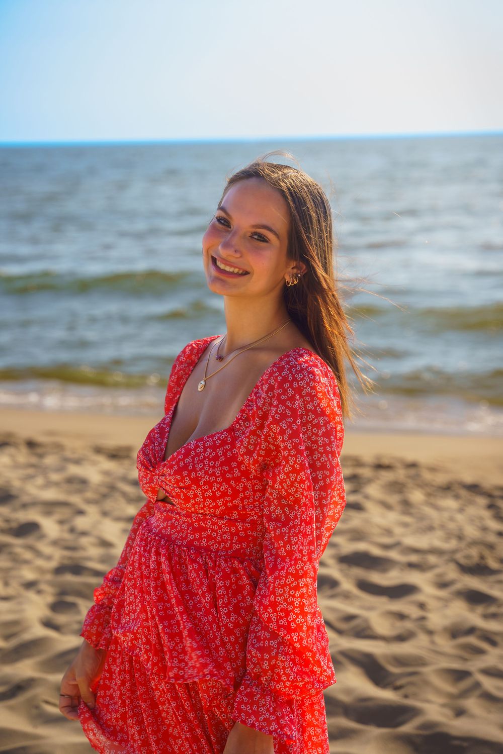 Smiling young woman wearing red floral long-sleeve dress standing on sandy beach by the ocean.
