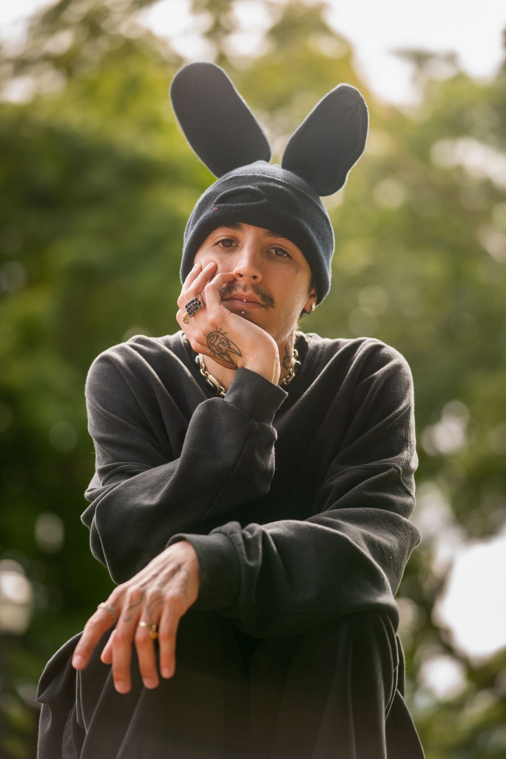 Young man in black hoodie with bunny ears beanie posing thoughtfully outdoors with green trees behind.