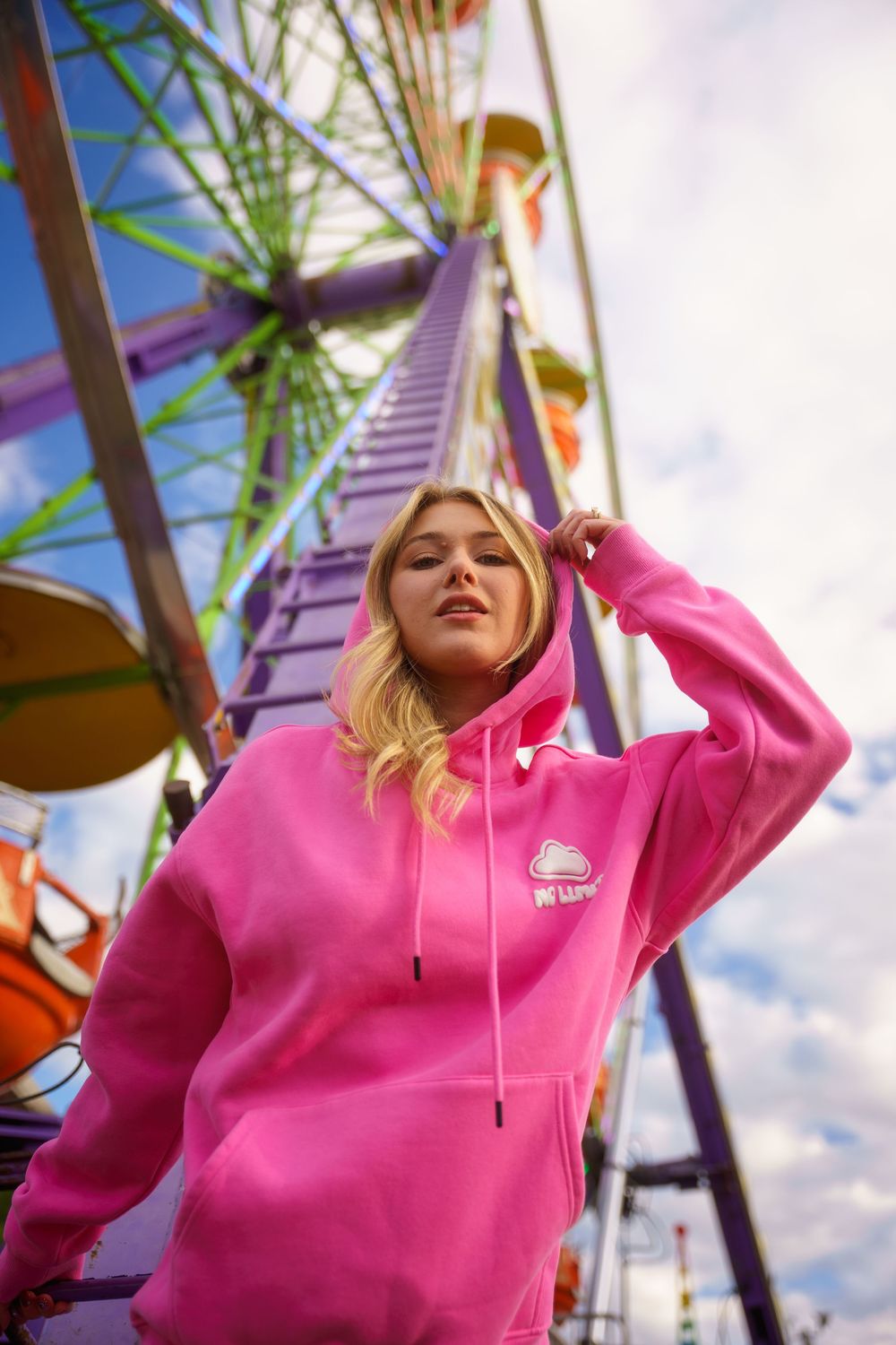 Blonde woman in bright pink hoodie posing in front of colorful Ferris wheel at amusement park.