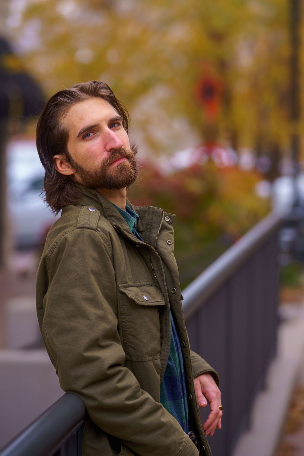 Bearded man in olive jacket leaning on railing with autumn foliage in urban background.