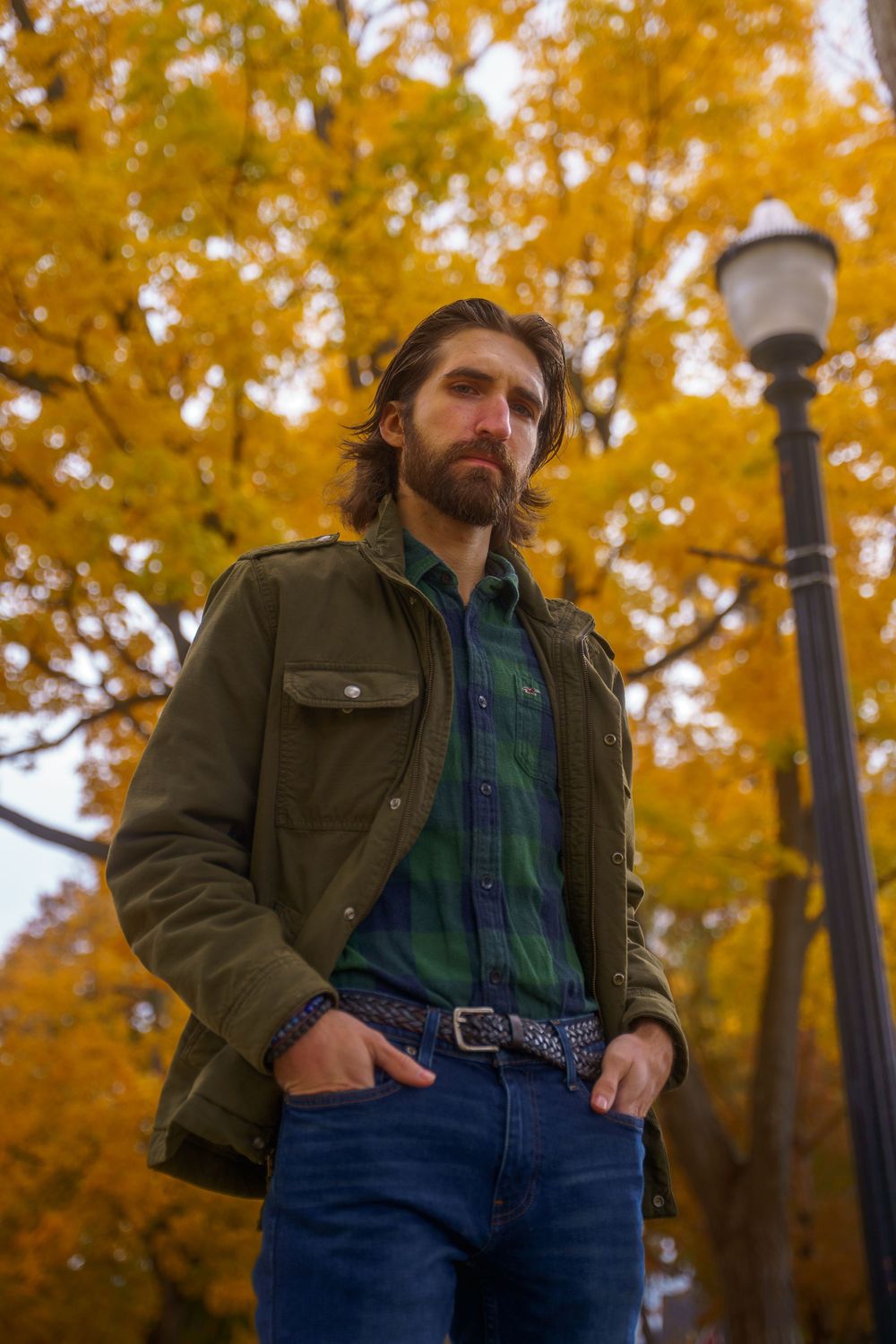 Bearded man in olive jacket and jeans standing before vibrant yellow autumn trees near lamppost.