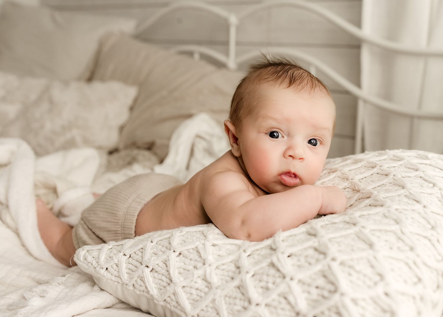 A chubby baby lies on a white knit blanket on a bed, propped up on their elbows, looking at the camera.