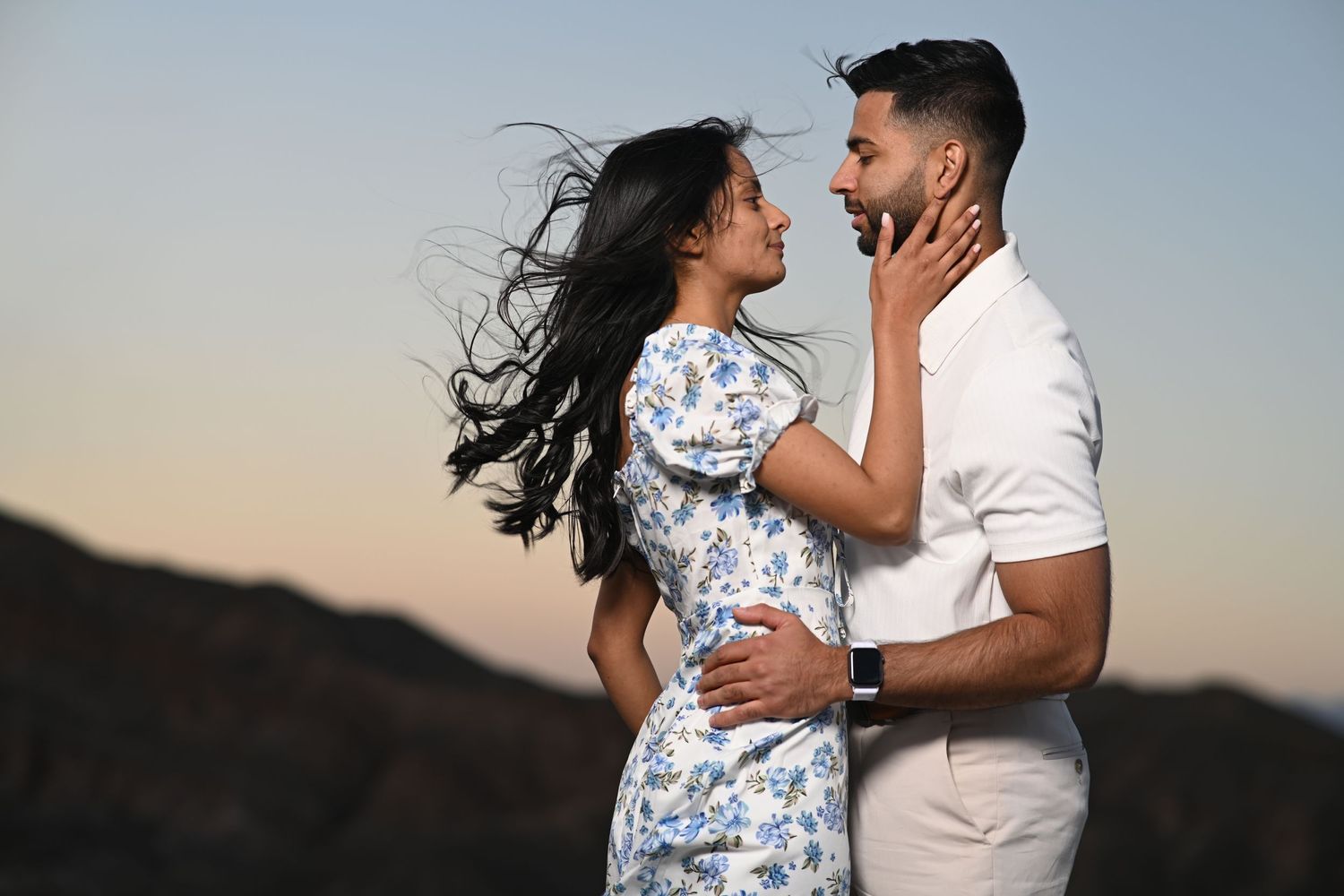 Couple embracing at sunset with wind-blown hair, sharing a romantic moment against a scenic mountain backdrop.