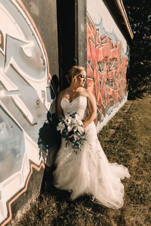 Bride in lace wedding gown poses against graffiti wall holding white and blue bouquet.