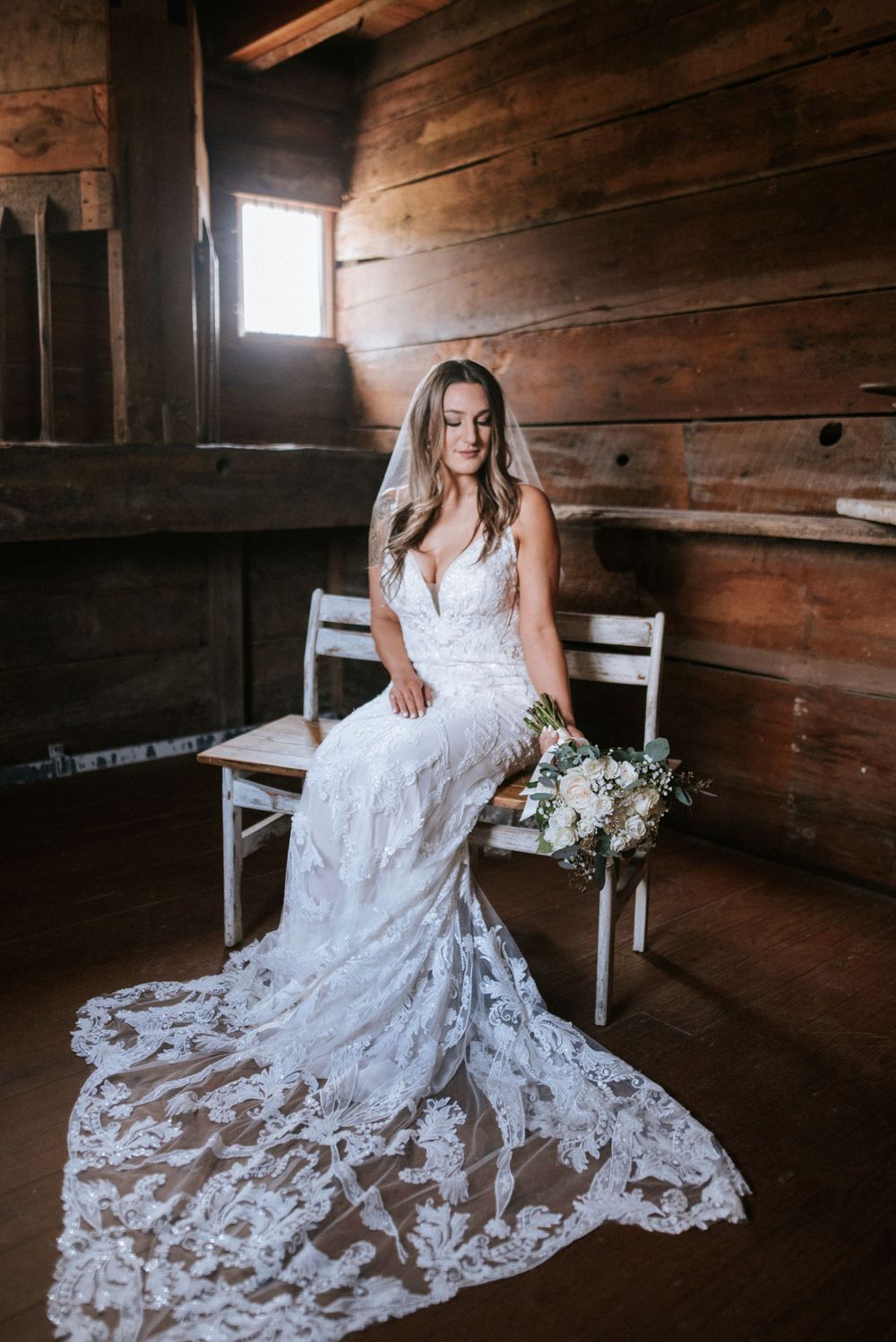 A white lace wedding dress with intricate details and long train photographed in rustic wooden barn setting with window light.