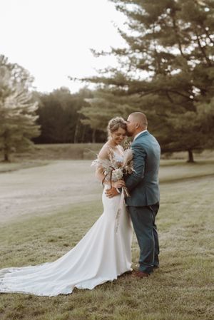 Newlyweds embrace on grassy hillside with pine trees in background.