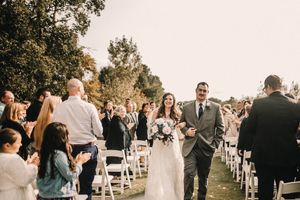Wedding guests line white chairs outdoors as a couple walks down the aisle after their ceremony.