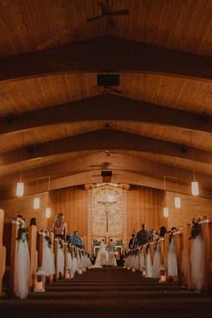 Wooden chapel interior with vaulted ceiling during wedding ceremony illuminated by pendant lights.
