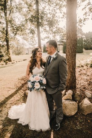 Outdoor wedding portrait with couple standing among tall trees with pastel floral bouquet.