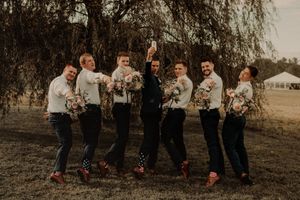 Groomsmen in suspenders pose playfully in field under willow tree at sunset.