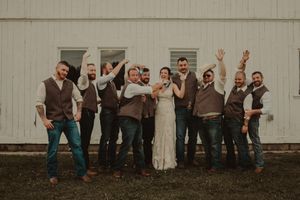 Wedding party wearing brown vests strikes fun pose against rustic white barn wall.