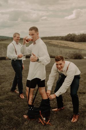 Groomsmen in suspenders helping put on shoes during wedding preparations outdoors.