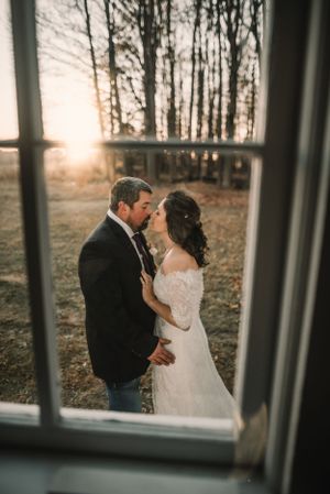 Romantic sunset kiss captured through window frame at rustic wedding venue.