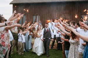 Wedding guests form a sparkler tunnel exit for the newlyweds outside a rustic wooden barn at dusk.