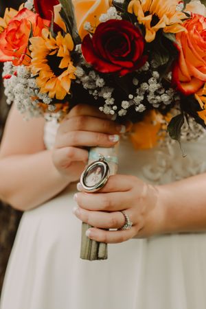Close up of bridal bouquet with orange roses, sunflowers and red roses held by hands with wedding rings.