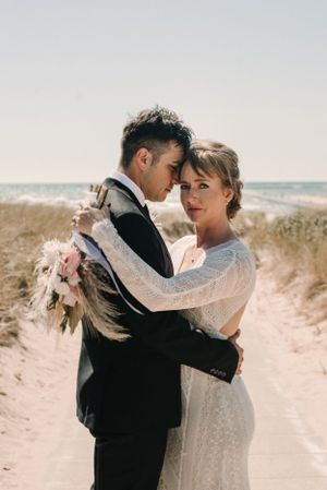 Beach wedding portrait with couple embracing against ocean backdrop and holding pampas grass bouquet.