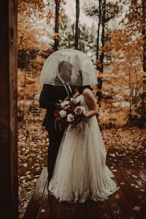 Newlyweds share an intimate moment under a clear umbrella surrounded by autumn foliage in the woods.
