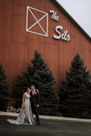 A wedding couple poses in front of The Silo venue with evergreen trees framing the rustic backdrop.