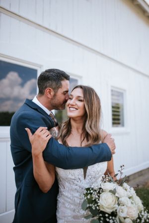 A joyful wedding couple embracing against a white barn wall with a bridal bouquet.