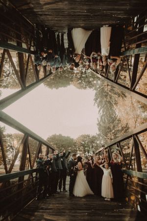 Wedding party celebrating on a covered wooden bridge with raised glasses.