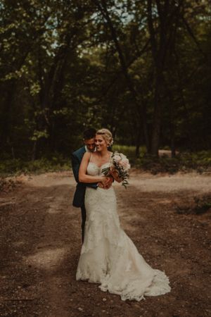 A romantic wedding photo of a couple embracing on a wooded path with the bride holding a bouquet.