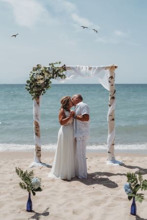 Beach wedding ceremony setup with birch arch decorated with white fabric and flowers as seabirds fly overhead.