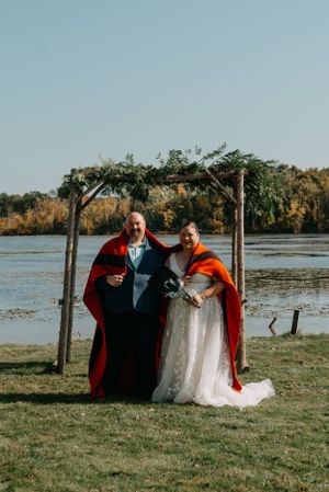 Couple in wedding attire and red capes pose under floral arch by waterfront.