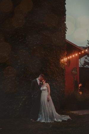 A romantic evening wedding portrait with glowing string lights and a red barn in the background.