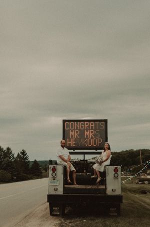 A couple sits atop a vintage truck with a congratulatory wedding sign under moody skies.