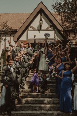 Wedding party and guests celebrate on church steps with raised arms and joyful expressions.