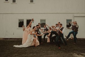 Wedding party poses playfully outside a white barn with synchronized jumping motion.