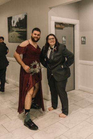 Two wedding guests pose playfully in formal attire, one wearing a burgundy dress and the other in a black suit.