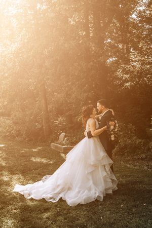 A romantic sunset wedding photo showing a couple embracing in a forest with the bride wearing a flowing layered gown.