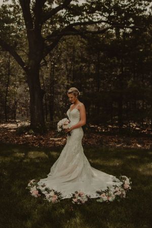 A bride in a flowing lace gown stands in a forest setting with flower petals arranged in a circle on the ground.