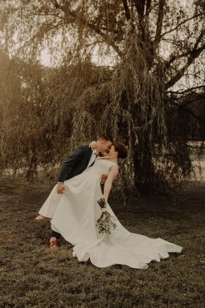 Wedding couple embraces romantically under a willow tree in a dreamy sepia-toned photograph.
