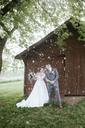 Newlyweds share a joyful moment in falling snow by a rustic barn.