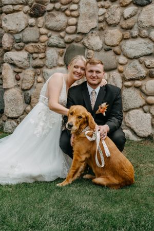 A wedding portrait with a couple and their golden retriever against a stone wall backdrop.
