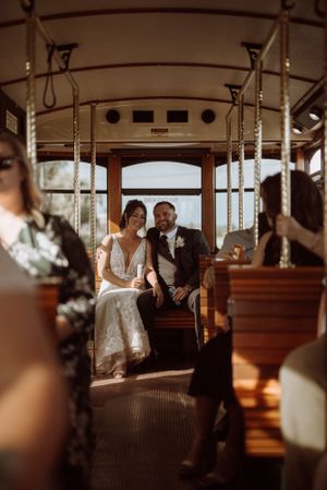 A couple sits together in a vintage trolley car on their wedding day with wooden benches and pole handles visible.