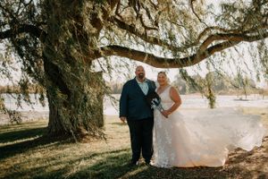 A couple poses under a large willow tree in their wedding attire on a sunny afternoon.