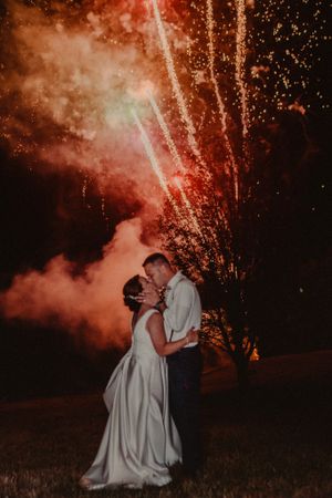 A dramatic wedding kiss under red fireworks in the night sky.