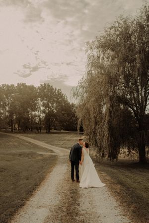 A couple in wedding attire walks down a serene path under a weeping willow tree at sunset.