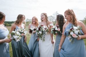 Bridesmaids in dusty blue dresses laughing together while holding floral bouquets.