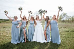 Wedding party in light blue dresses poses with bouquets in sunny field.