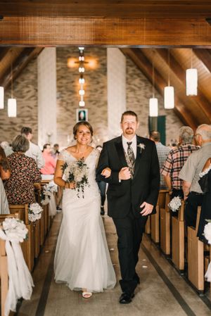 A wedding couple walks down the aisle of a church with wooden beams and pendant lights.