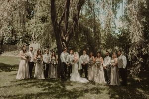 Wedding party poses for a group photo under willow trees in natural outdoor lighting.
