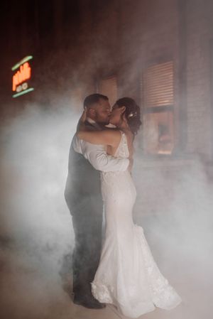 Romantic couple shares first dance in atmospheric fog with neon sign glowing in background.